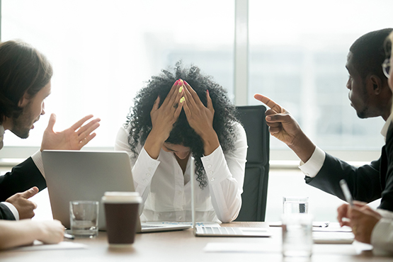 Woman sitting at a desk with her head in her hands being shouted at by a colleague